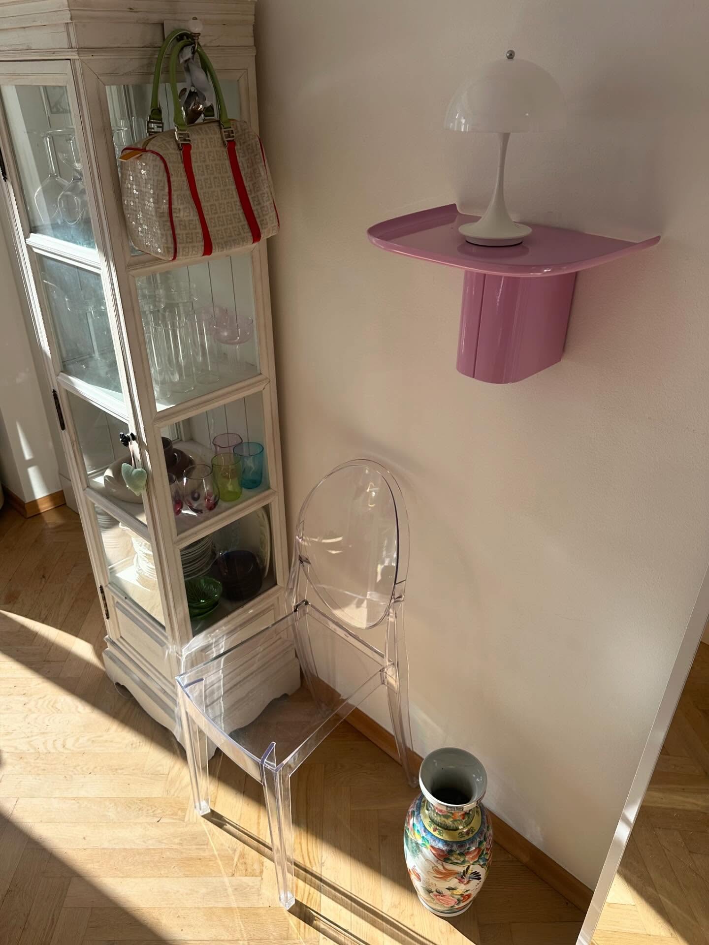 Transparent plastic chair against wall, colorful vase on floor, pink wall-mounted shelf with white table lamp, glass cabinet displaying porcelain and glasses.