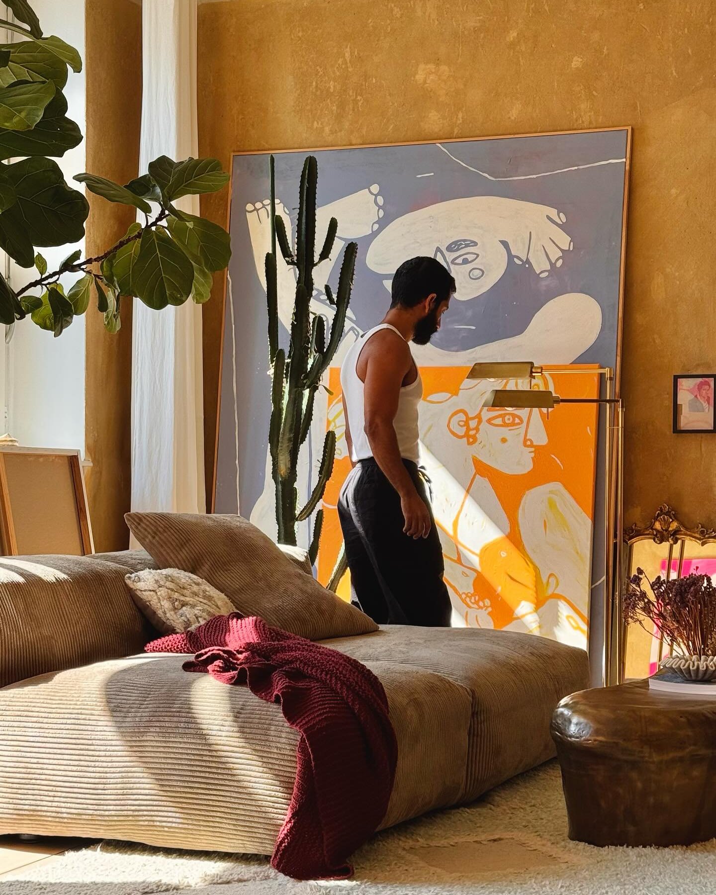 Cozy living room with a corduroy beige sofa, brown cushions, a red knitted blanket, large colorful abstract paintings, a tall cactus plant, and a leather ottoman with books.