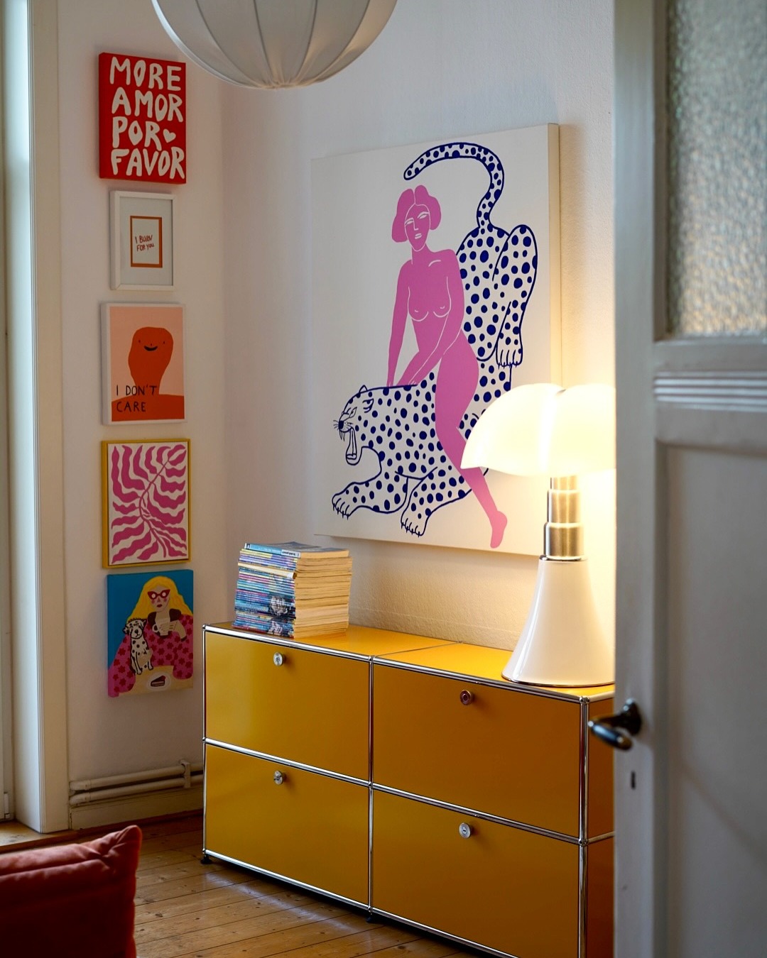 Yellow sideboard with stack of CDs and modern white table lamp beneath colorful wall art.