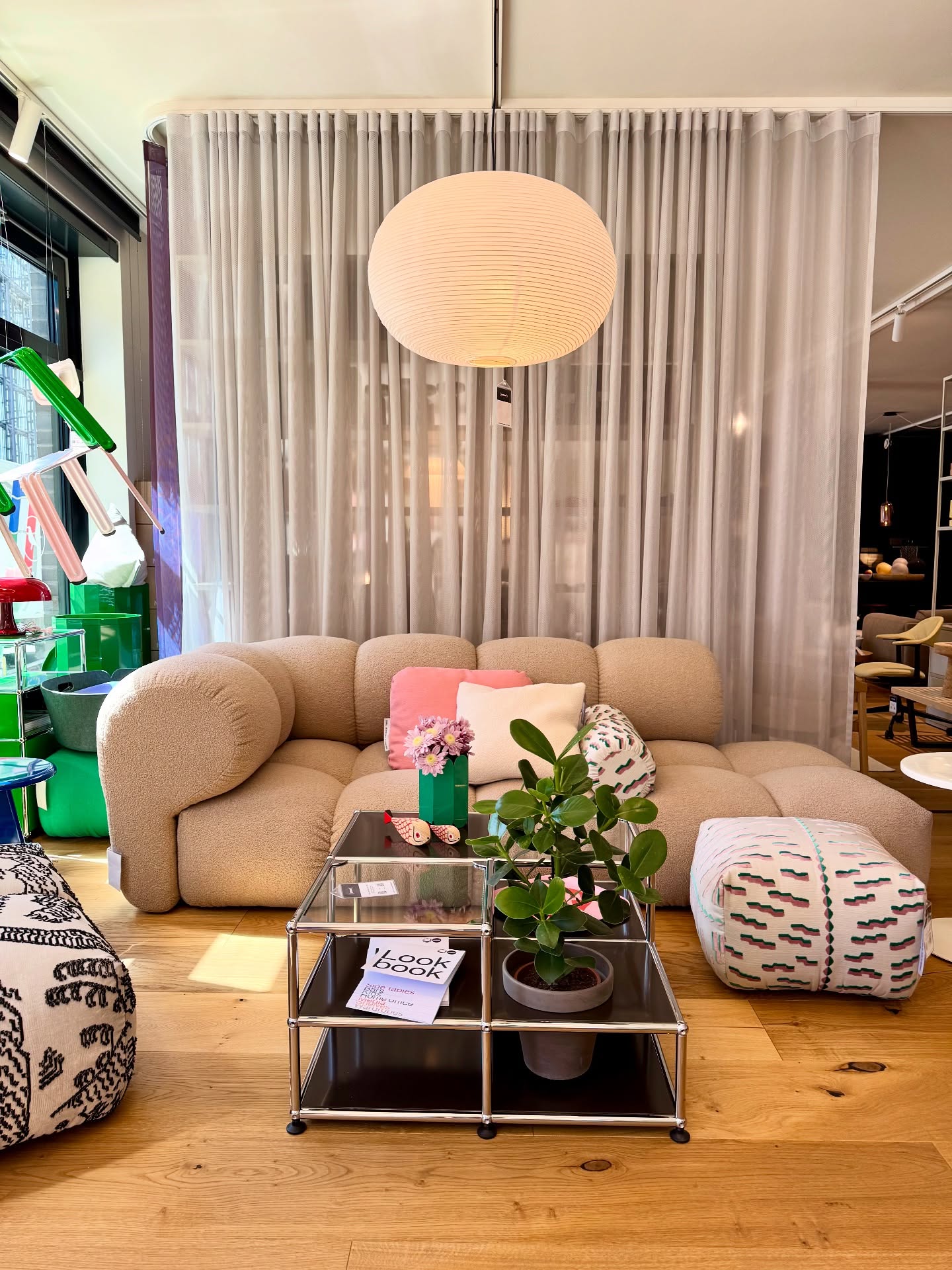 Metal and glass coffee table with two green plants, a green vase with purple flowers, and a book, in front of a beige sofa with colorful cushions.