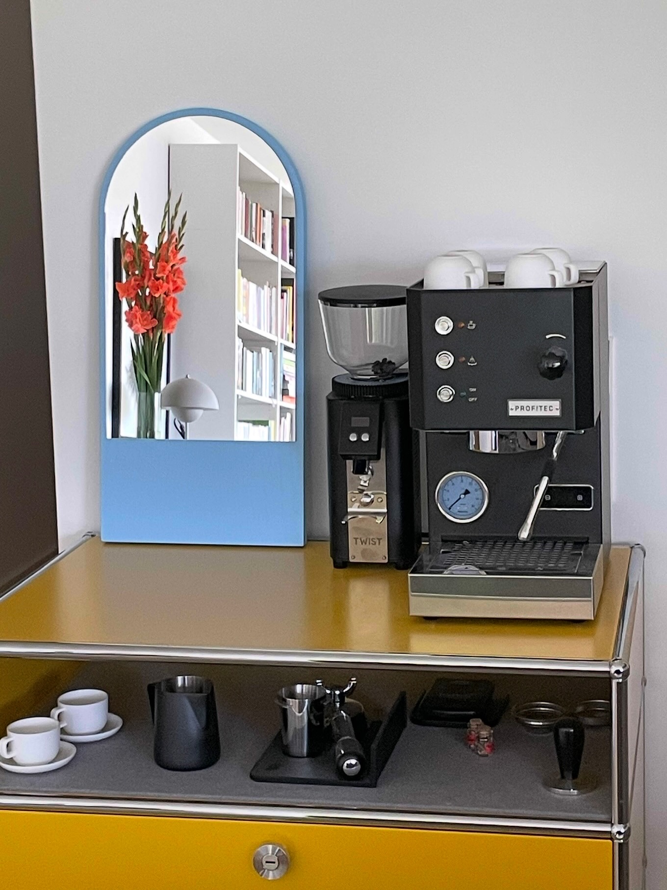 Coffee machine on yellow cabinet next to yellow lamp in modern kitchen with white countertops and citrus fruits in the foreground.