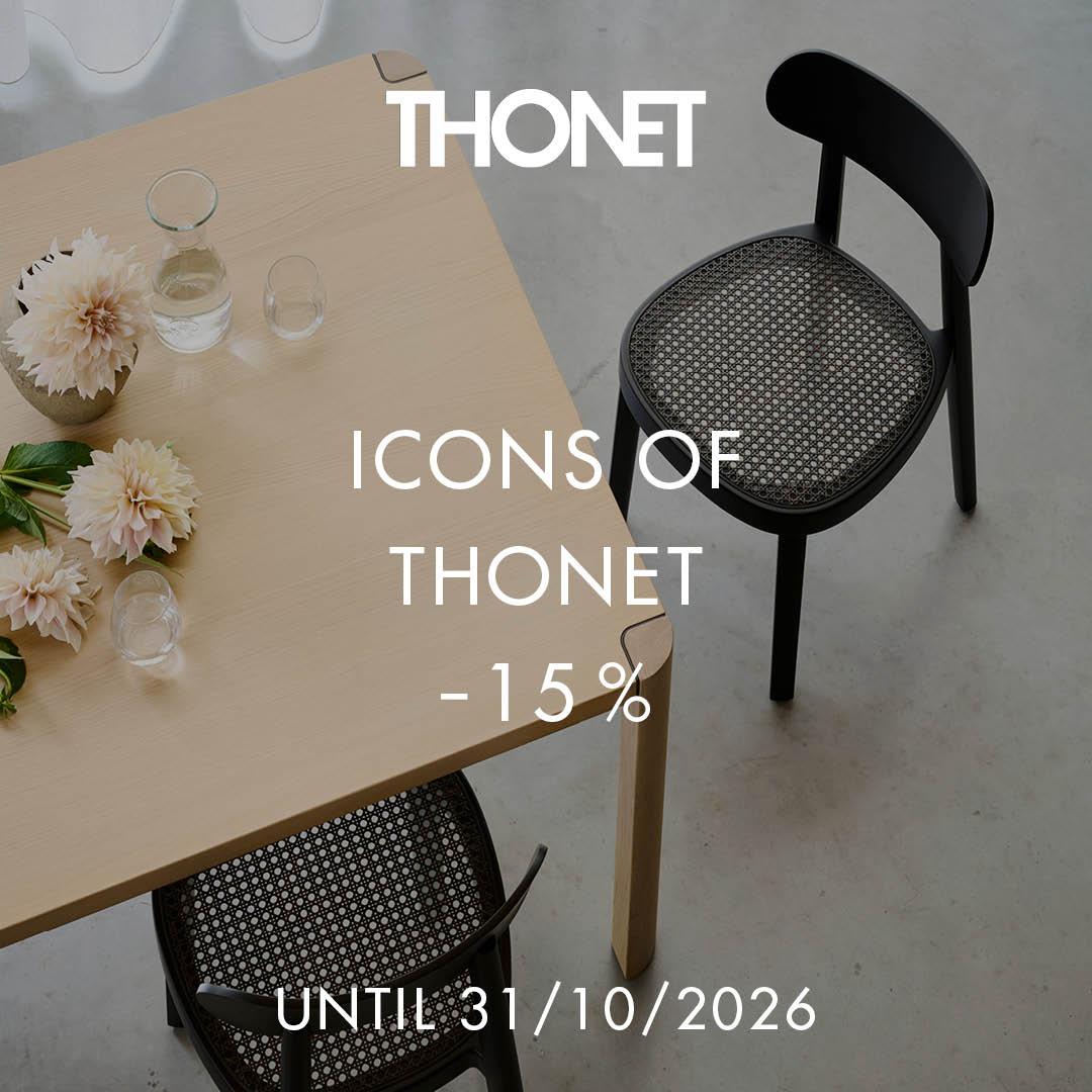 Light wood table with minimalist design, decorated with flowers and glassware, paired with black chairs featuring woven cane seats, on a concrete floor.